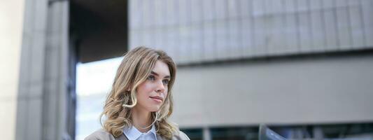 Vertical shot of young confident woman in suit, holding her laptop and smiling, being outside on a street, going to work photo