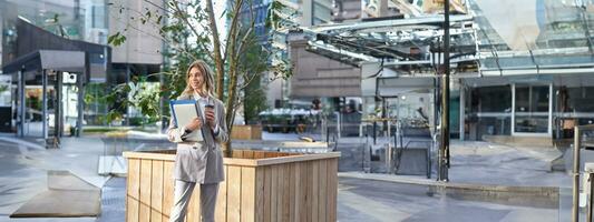 elegante corporativo mujer en traje, en pie en calle con documentos, trabajo ordenador portátil y café, esperando cerca negocio centrar foto