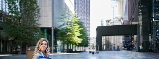 Vertical shot of young woman sitting near office building, holding laptop and folders with work documents, calling someone on mobile phone photo