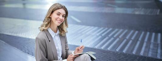 Business woman in suit sits in city centre, writes down, takes notes, holds pen and notebook, brainstorms, creates ideas photo