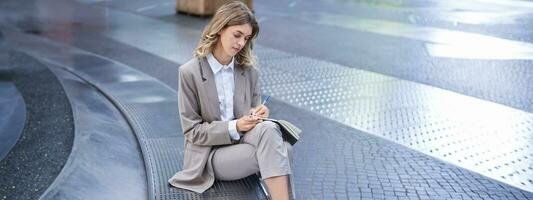 Business woman sits in city centre and works on mobile phone, she has her laptop and wears working corporate outfit photo