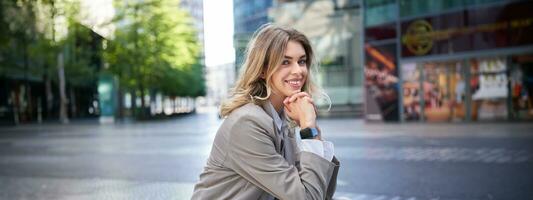 Portrait of woman waiting for an interview, sitting in city center in beige suit, has digital watch, smiling at camera photo