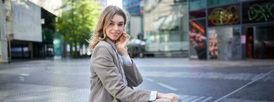 Confident blond businesswoman in suit, smiles at camera, sits outdoors near office buildings photo