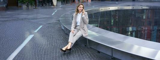 Young female manager. Corporate woman sitting outside on street and talking on mobile phone. Girl making phone call photo