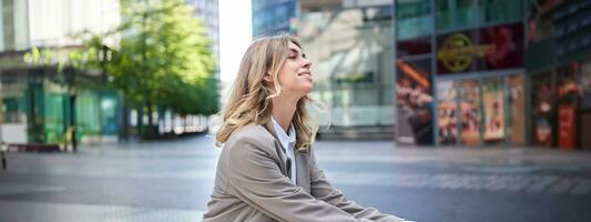 Smiling happy businesswoman in suit, sitting relaxed in city center and enjoying being outdoors photo