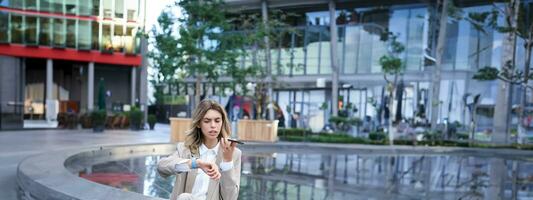 Vertical shot of smiling woman records voice message and looks at digital watch, reads message or checks time, sits in city centre photo