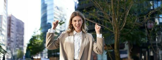 Excited businesswoman screams and celebrates, lifts hands up, does hooray gesture, celebrates victory, achieve goal, stands on street photo