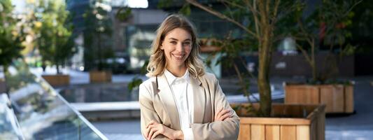 cerca arriba retrato de confidente y exitoso mujer de negocios en traje, cruzar brazos en pecho, en pie en poder actitud en calle cerca oficina edificio foto