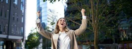 Happy businesswoman scream of joy, raise hands up in celebration, cheering gesture, achieve goal and triumphing on city street photo