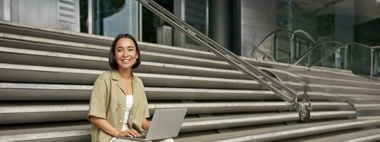 Vertical shot of asian girl sits with laptop, drinks coffee on university stairs. Young woman, student does her homework outdoors photo