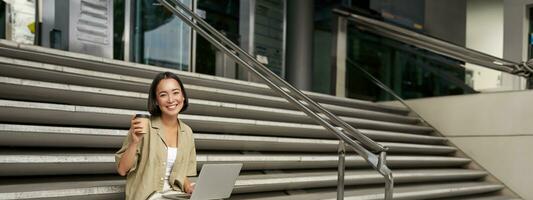 Vertical shot of smiling girl student, asian woman sits on stairs of university campus and drinks coffee, does her homework on laptop photo