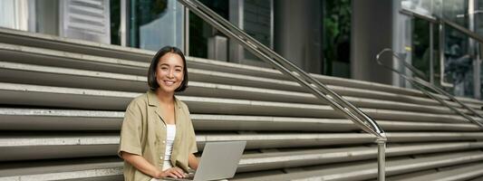 Vertical shot of asian girl sits with laptop, drinks coffee on university stairs. Young woman, student does her homework outdoors photo