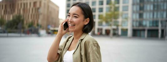 Modern young asian girl talks on mobile phone, uses telephone on city street. Woman smiling while calling someone on smartphone photo