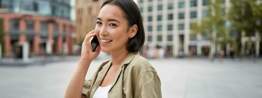 celular conexión. joven asiático mujer hace un teléfono llamar, hablando en móvil teléfono inteligente y caminando en calle foto