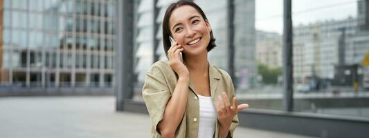 Cellular connection. Young asian woman makes a telephone call, talking on mobile smartphone and walking on street photo