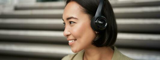 Close up portrait of smiling asian girl in headphones, listens to music outdoors, looking happy. photo