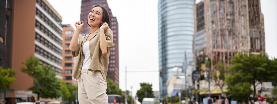 vertical Disparo de joven asiático mujer posando feliz, levantamiento manos arriba y baile, triunfando, celebrando victoria, disfrutando día fuera en ciudad foto