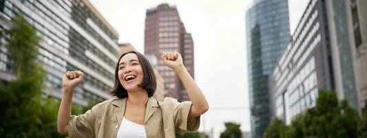 Happy people. Young smiling asian woman dancing in city, raising hands up in city, triumphing, celebrating victory photo