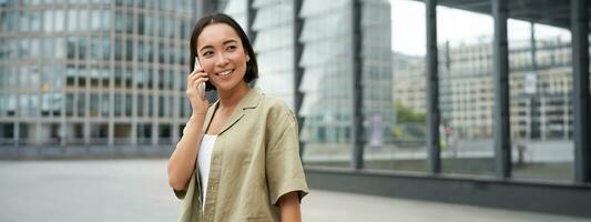 Portrait of asian girl smiling, talking on phone, making a call, standing on street near building and waiting for someone, answer telephone photo