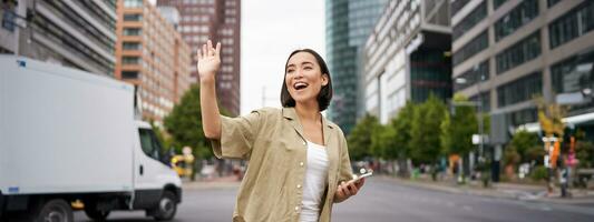 Happy asian girl passing by friend and waving at them on street, saying hello while walking in city, holding smartphone photo