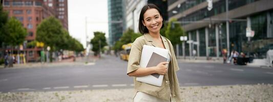 Portrait of young asian woman, looking happy and confident, going to work or university, city skyscrappers behind her, holding laptop and notebook photo