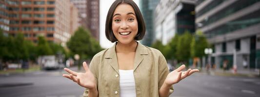 Modern asian girl standing on street with surprised face, looking impressed and amazed, hear awesome news photo