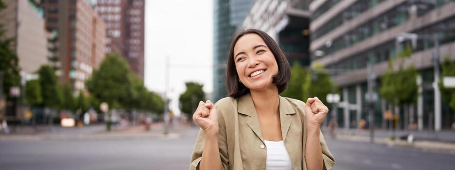 contento asiático niña triunfando en calles de ciudad, bailando desde felicidad, celebrando victoria, posando al aire libre foto