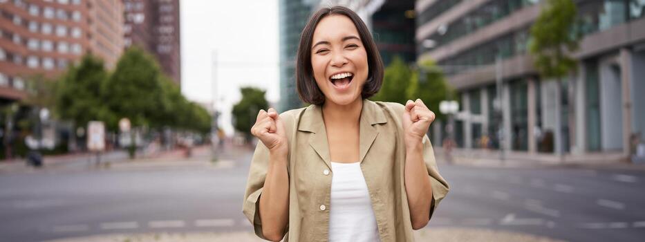 contento asiático niña triunfando en calles de ciudad, bailando desde felicidad, celebrando victoria, posando al aire libre foto