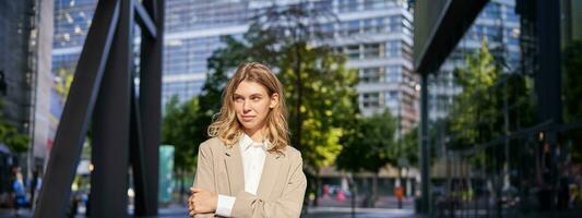 Portrait of young saleswoman, confident businesswoman in suit, cross arms on chest, standing in power pose on street near office buildings photo