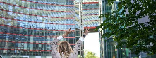 Corporate people. Rear view of businesswoman raising hands up in celebration, triumphing, achieve goal and rejoicing, standing on street photo