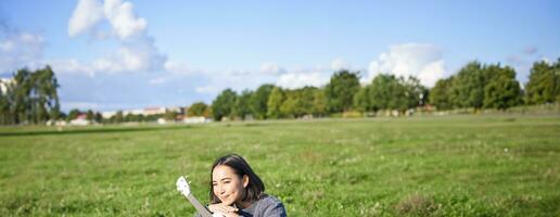 Romantic asian girl sitting with ukulele guitar in park and smiling, relaxing after university, enjoying day off on fresh air photo