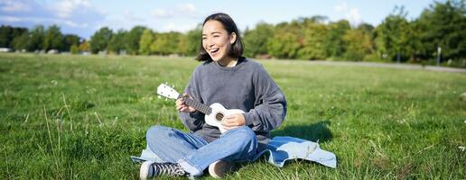 contento morena asiático chica, estudiante jugando ukelele sentado en césped en parque, relajante, canto canción, estilo de vida concepto foto