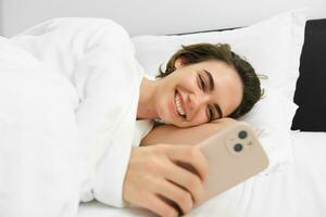 Image of woman lying in bed with white linen sheets, looking at smartphone and laughing, watching funny video on mobile phone app photo