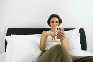 Portrait of young modern woman lying in her bed and using mobile phone, resting in bedroom and looking at smartphone screen photo