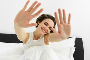 Portrait of sleepy, happy young woman, wakes up in her bed, stretching out hands, hiding face from camera, smiling and laughing photo