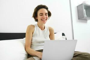 Modern woman resting at home, sitting on bed with laptop, looking at computer screen, laughing and smiling photo