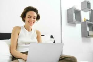 Portrait of young beautiful woman working from home, online shopping on laptop, using her computer and sitting on bed, resting in bedroom photo