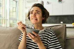 Close up portrait of female model, girl eating breakfast and watching intense scene in movie, staring at tv screen in living room, holding bowl of cereals with milk photo