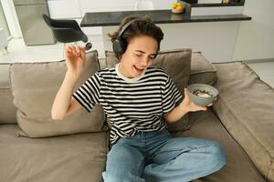 retrato de elegante joven mujer escuchando a música en auriculares, comiendo cereales con Leche en sofá en vivo habitación, bailando mientras sentado y teniendo desayuno, participación cuenco y cuchara foto