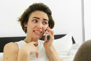Image of woman with anxious, awkward face expression, talking on mobile phone, cringe from something she hears during conversation, lying in bed, saying something risky photo