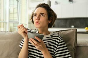 Portrait of girl eating bowl of cereals and watching movie on tv, looking at screen with serious face, having breakfast in living room photo