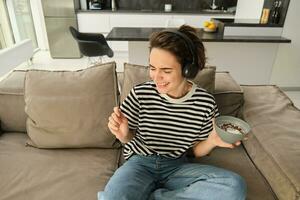 retrato de despreocupado sonriente mujer, escucha música en auriculares, participación cuenco con cereales, comiendo desayuno, sentado en sofá en vivo habitación y haciendo danza se mueve foto