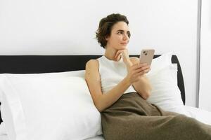 Woman looking with thinking face, using phone, lying in bed in white tank top and trousers photo