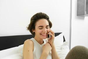 Female model sitting on bed with phone, calling friend, chatting over telephone, laughing and smiling photo
