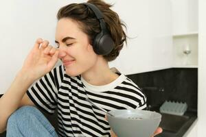 retrato de despreocupado mujer en auriculares, comiendo su desayuno, participación cuenco de cereales, riendo y sonriente, sentado en el cocina en un mostrador foto