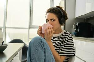 Close up portrait of smiling brunette woman, student drinks her tea and listens music or ebook in headphones, sits in kitchen and relaxes photo