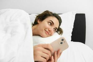 Portrait of woman lying in her bed, checking phone, sets up alarm clock to wake up in the morning photo