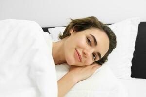 Close up portrait of smiling young woman, relaxing, lying in bed with calm, tender face expression, resting, going to sleep photo