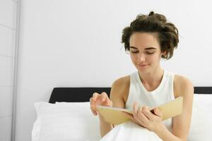 Portrait of beautiful smiling woman, flipping notebook pages, reading journal, going to sleep in bed and revising for exam photo