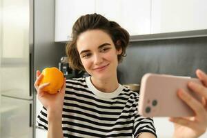 Smiling, beautiful young woman taking selfie with fresh yellow pepper, food blogger taking selfie with vegetables in the kitchen, vegan making a meal and recording video for social media photo